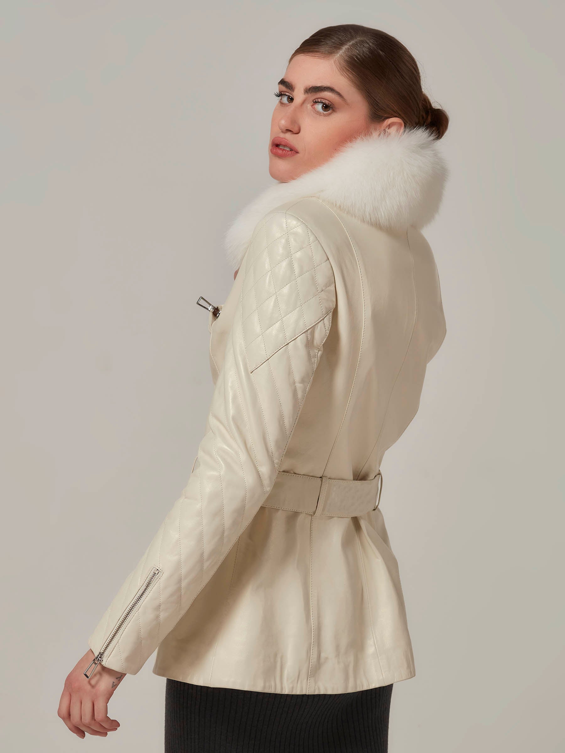 A woman wearing a white leather motorcycle jacket with a luxurious white fox fur collar, featuring quilted details on the shoulders. Side pose.