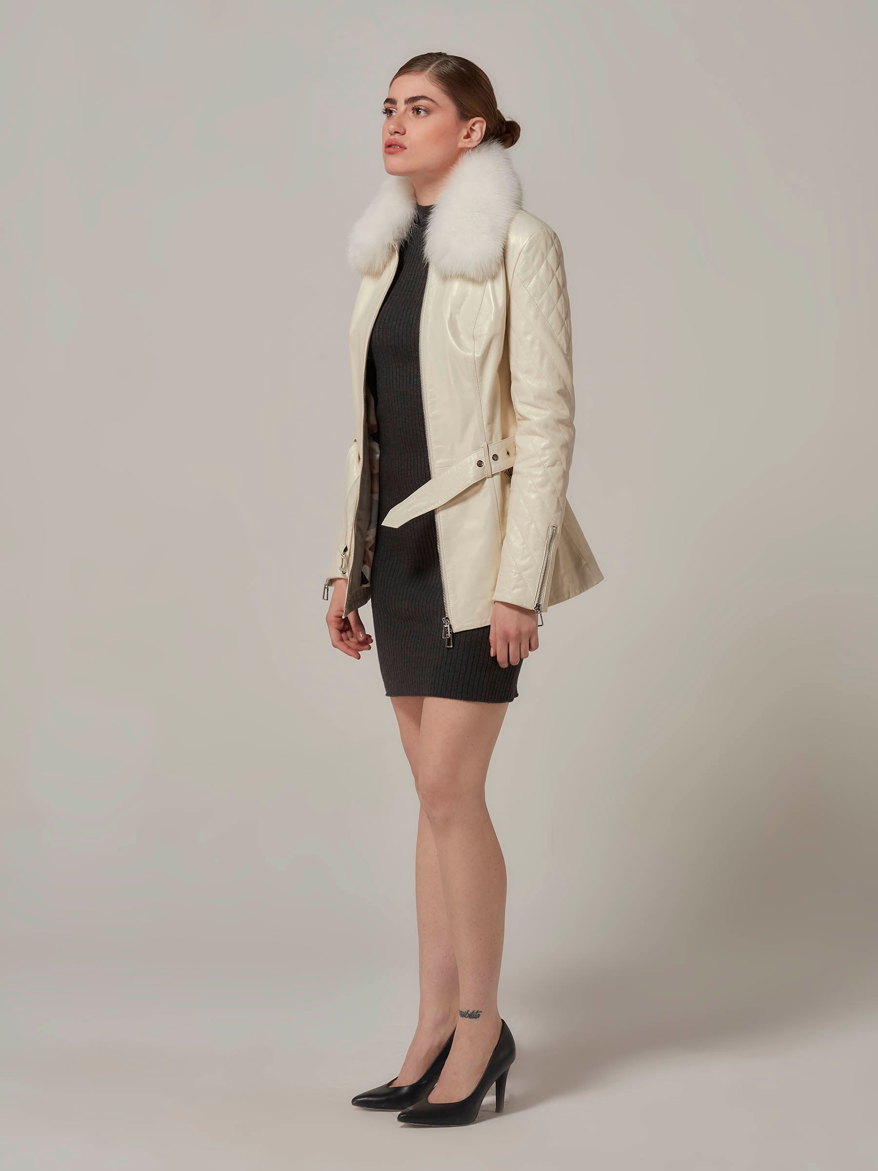 A woman wearing a white leather motorcycle jacket with a luxurious white fox fur collar, featuring quilted details on the shoulders. Side full length pose.